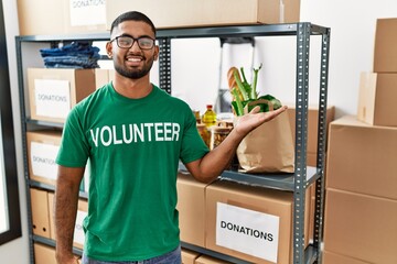 Sticker - Young indian man volunteer holding donations box smiling cheerful presenting and pointing with palm of hand looking at the camera.