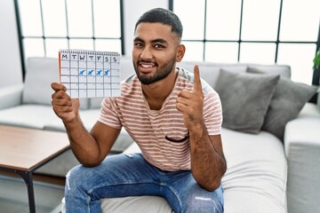 Wall Mural - Young indian man holding travel calendar smiling with an idea or question pointing finger with happy face, number one