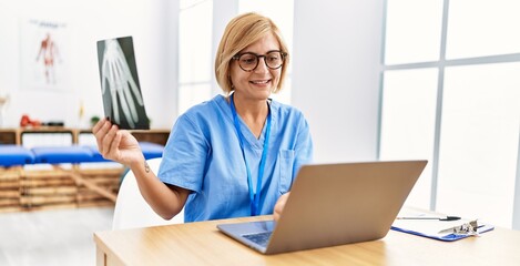 Sticker - Middle age blonde woman wearing physio therapy uniform holding xray using laptop at clinic