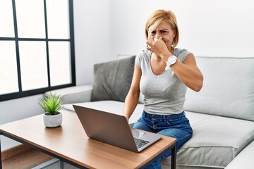 Middle age blonde woman using laptop at home smelling something stinky and disgusting, intolerable smell, holding breath with fingers on nose. bad smell