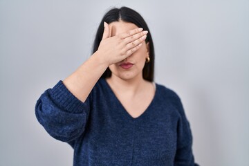 Sticker - Young brunette woman standing over isolated background covering eyes with hand, looking serious and sad. sightless, hiding and rejection concept