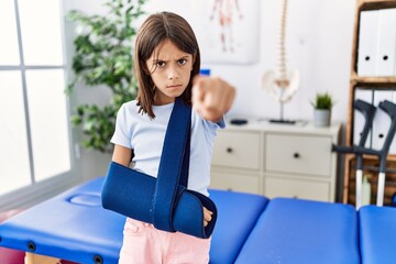 Poster - Young hispanic girl wearing arm on sling at rehabilitation clinic pointing with finger to the camera and to you, confident gesture looking serious