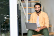 © insta_photos - Smiling ethnic indian man wearing earphones sitting at home on stairs using laptop computer looking at camera. Remote work or elearning online concept. Hybrid virtual chat video calls. Portrait
