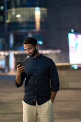 Wall Mural - Indian business man wearing earbuds standing outdoors on night big city street holding cellphone using smartphone looking at mobile phone watching streaming tv game or having mobile chat video call.