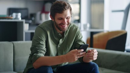 joyful man is chatting online by smartphone sitting in living room, portrait of smiling guy in home