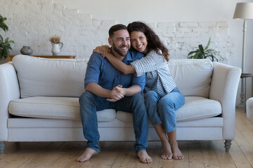 Wall Mural - Multi ethnic attractive couple in love hugging seated on sofa at home laughing enjoy dating, spend carefree weekend together. Homeowners family portrait, relationships, happy marriage, tenancy concept