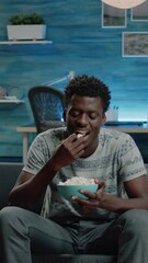 Wall Mural - Vertical video: Black adult watching comedy movie on television while eating popcorn from bowl. African american person laughing and looking at TV with snack. Man of african ethnicity having fun