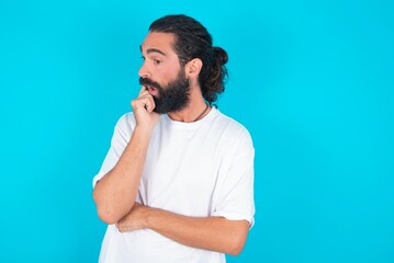 Wall Mural - Astonished young bearded man wearing white T-shirt over blue studio background looks aside surprisingly with opened mouth.