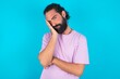 © Roquillo - Very bored young bearded man wearing violet T-shirt over blue studio background holding hand on cheek while support it with another crossed hand, looking tired and sick,