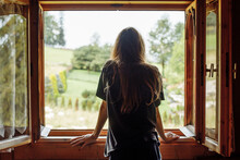 Rearview Of Long Dark-haired Woman In T Shirt Looking Out The Window, Contemplating Landscape Rustic Natural Tree View