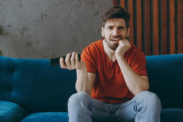 Wall Mural - Young sad man wears red t-shirt hold TV remote controller watch film switch channels sit on blue sofa couch stay at home hotel flat rest relax spend free spare time in living room indoors grey wall.