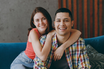 Young smiling happy fun cool couple two friends family man woman in casual clothes look camera sits on blue sofa together stay at home flat rest spend free spare time in living room indoors grey wall.