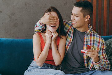 Young couple two friends family man woman in casual clothes makes a wedding proposal cover eyes sit on blue sofa together stay at home flat rest spend free spare time in living room indoor grey wall