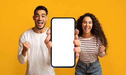 Wall Mural - Happy emotional millennial arab couple enjoy online victory, show big smartphone with empty screen