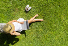 Woman In A White Dress, Hat With Laptop, Sitting On The Green Grass Under Tree At Summer Day. Top View, Drone, Aerial View