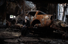 A Girl In A Black Suit And A White Mask Sits Near A Burnt-out Car From A Russian Army Shell Hitting The House.
