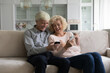 © fizkes - Happy cheerful retired couple using online apps on smartphones, talking, discussing social media content, holding mobile phones, resting on home sofa, hugging, smiling, laughing. Communication