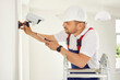 © Studio Romantic - Professional young electrician worker in uniform and cap installs and screws surveillance video camera on wall in bright office space, school, hospital. He is standing on stepleader. Repair works.