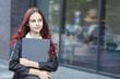 © upparadox - Young girl with closed laptop standing in front of a glass window in the city