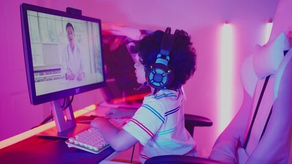Cute little curly-haired Asian boy in a gaming headset having video online chatting with Asian male doctor by using the application for video call on a personal computer in a violet neon room at home.