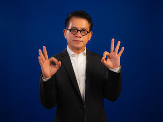 Portrait of smart middle-aged asian confident successful businessman wearing suit showing okay sign on blue background in studio, looking attractive and positive professional leadership