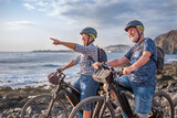 Senior caucasian couple riding off road on the pebble beach with bicycles at sunset looking away smiling. Authentic elderly retired life concept