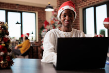 Portrait Of African American Employee Wearing Santa Hat, Working On Business At Company Office Filled With Christmas Decorations And Tree Lights. Using Laptop In Startup Workplace With Xmas Decor.