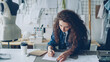 © silverkblack - Young female artist fashion designer is drawing women's garment sketch with pencil at table in modern studio. Mannequin, sewing items, take-away coffee are visible.