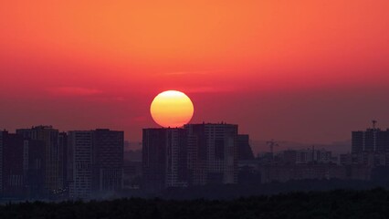 Wall Mural - A big red sun in the sunset sky over the roofs of buildings, urban landscape. Evening sky in bright sunlight over the twilight city