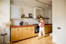 Stylish Kitchen Interior Of Modern Apartment With Motion Blurred Female Person Walking Inside. Interior Made In White And Beige Tones With Wooden Kitchen Front And Floor