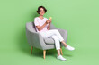 © deagreez - Full size photo of cute young woman sit armchair relaxing hold device typing dressed trendy striped look isolated on green color background