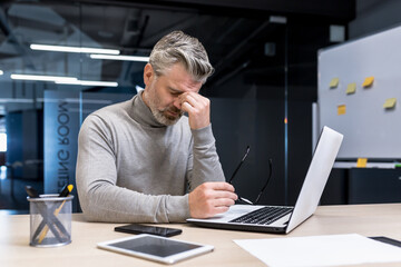 Senior gray haired businessman overtired working on laptop, mature man wearing glasses has pain in eyes, boss working in company inside modern office building.