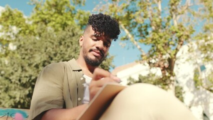 Young male student wearing an olive colored shirt takes notes in notebook while sitting on bench outside the university