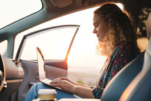 Beautiful Curly-haired Woman Working With Laptop Sitting On The Front Seat Of A Car On A Sunny Day. Remotely Relaxing. Leisure, Travel, Tourism. Road Trip.