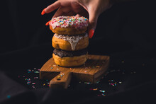 Close-up Of A Woman's Hand Taking A Delicious Donut From A Wooden Board. Delicious Donuts Laid Out On A Wooden Cutting Board On A Black Background