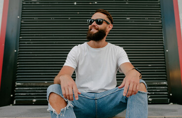 Wall Mural - Young bearded hipster guy wearing white blank t-shirt and blue jeans in a random urban style. Design men t shirt mock-up for print
