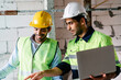 © thanmano - Two construction engineers in vest with wear safety hard hat hold laptop computer and talk about construction work on building site