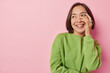 © WHstudio Leushin N - Happy millennial girl with dark hair covers face with hand smiles joyfully looks aside feels carefree dressed in casual green jumper isolated over pink background empty space for your promotion