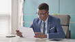 © stockbakers - Young African Businessman Reading Reports while Sitting in Office