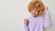 Excited Happy Woman With Curly Bushy Hair Makes Fist Pump Exclaims Yes Celebrates Win And Goals Achievement Dressed Inn Casual Purple Jumper Looks Aside Isolated Over Grey Background Blank Space