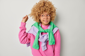 Wall Mural - Photo of lovely woman curls hair being deep in thoughts concentrated away considers something wears spectacles casual pink jumper pullovers tied over shoulders isolated over grey background.