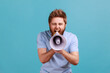 Portrait of bearded man holding megaphone near mouth, loudly speaking, screaming, making announcement, paying attention at social problems. Indoor studio shot isolated on blue background.