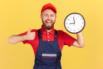 Wall Mural - Portrait of happy smiling handyman standing with big wall clock in his hands, looking at camera and showing thumb up, wearing overalls and red cap. Indoor studio shot isolated on yellow background.