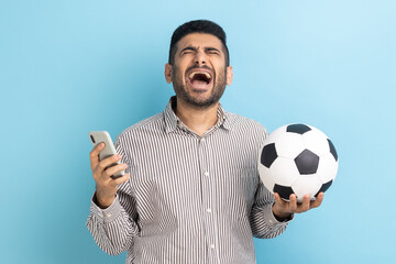 Portrait excited bearded businessman holding smart phone, betting and winning, screaming happily, celebrating, wearing striped shirt. Indoor studio shot isolated on blue background.