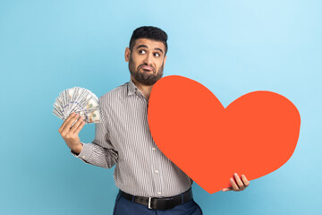 Portrait of doubtful confused bearded businessman standing with uncertain expression, holding fan of money and big red heart, wearing striped shirt. Indoor studio shot isolated on blue background.