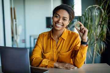 Poster - Excited black businesswoman holding golden bitcoin and smiling at camera, sitting at desk with laptop in office