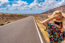 Back View Of Hipster Girl Wanderlust Walking On Asphalt Road In Wild Lands On Fuerteventura Island - Female Tourist With Hat Having Journey In Canary Islands Hitchhiking On The Way - Travel Concept