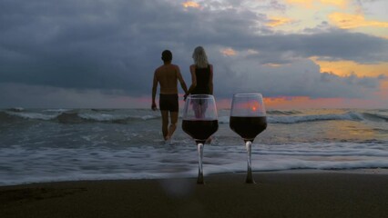 Wall Mural - Young woman man enters ocean or sea knee-deep against backdrop of beautiful sunset and glasses of red wine of rare plan. Honeymoon or romantic relationships of lovers. Love. Valentine's day.