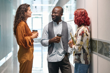 Leinwandbilder - Three young successful office managers in casualwear having chat while standing between two walls in corridor of modern building