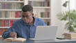 © stockbakers - Young African Man Reading Book and Working on Laptop in Office